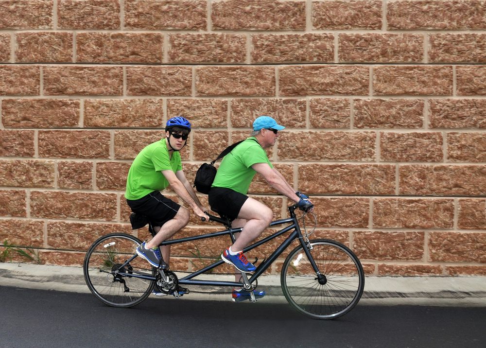 Two men wearing green shirts - riding their bikes with a brick wall behind them.