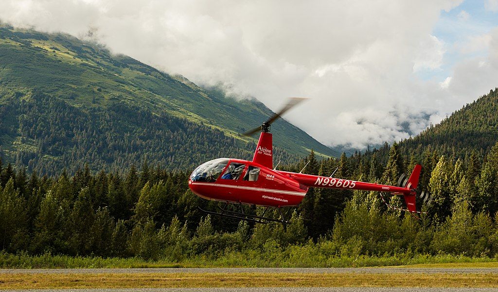 Red helicopter taking off near a field of trees.